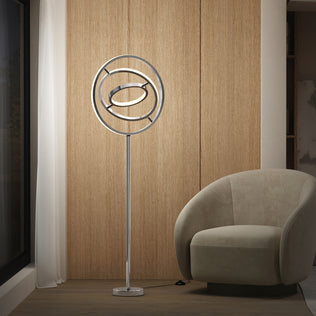 The Artika Pivot modern LED floor lamp 3 CCT, with a metallic circular frame, stands beside a beige curved armchair in a minimalist room featuring wood panel walls, a large window, and sheer white curtains.