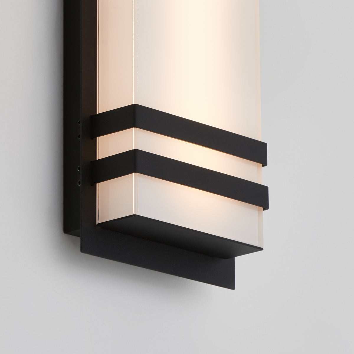A modern wall sconce with a frosted rectangular glass shade, accented by two horizontal black metal bands and a black base, mounted on a light-colored wall.