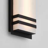 A modern wall sconce with a frosted rectangular glass shade, accented by two horizontal black metal bands and a black base, mounted on a light-colored wall.