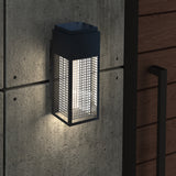 A modern outdoor wall sconce with a black metal frame and grid pattern emits a soft white light, mounted on a concrete wall next to a wooden door with a black handle.