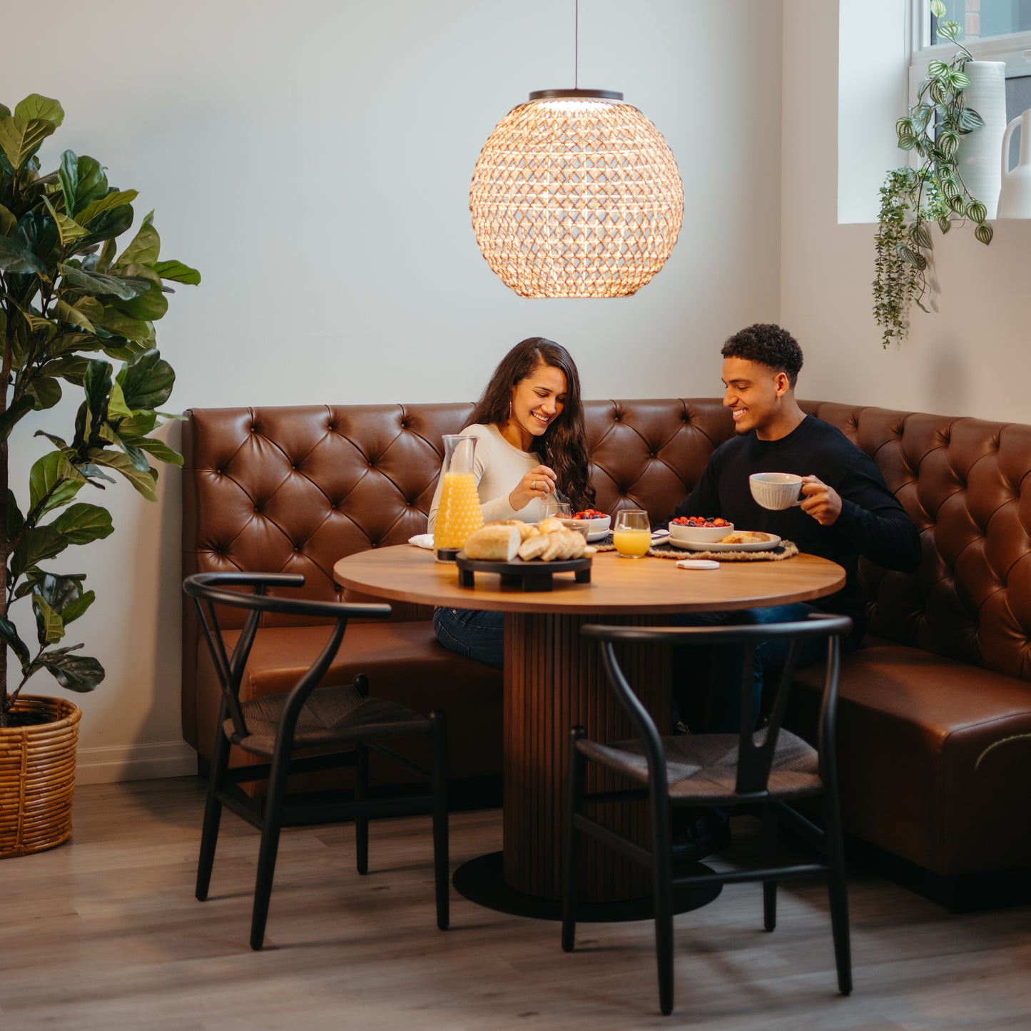 A smiling couple enjoys breakfast at a round table beneath the Benson LED pendant light by Artika, in a modern dining nook filled with warm lighting and plants for an inviting atmosphere.