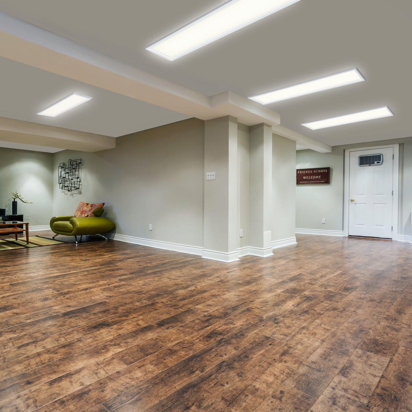 A spacious basement featuring Artikas Sunray ultra thin LED light panel (3 CCT), wood flooring, beige walls, a green chair, small coffee table, wall decor, and a Friends always welcome sign near a white door.
