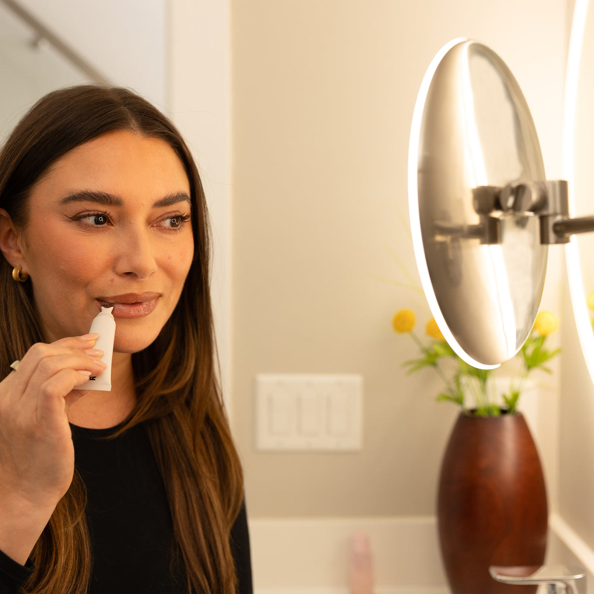 A woman with long brown hair uses a small white facial device on her upper lip while looking into a round, illuminated bathroom mirror. A vase with yellow flowers sits on the counter in the background.
