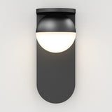 A modern wall-mounted light fixture with a matte black base and a half-sphere white diffuser, emitting soft white light against a plain white wall.
