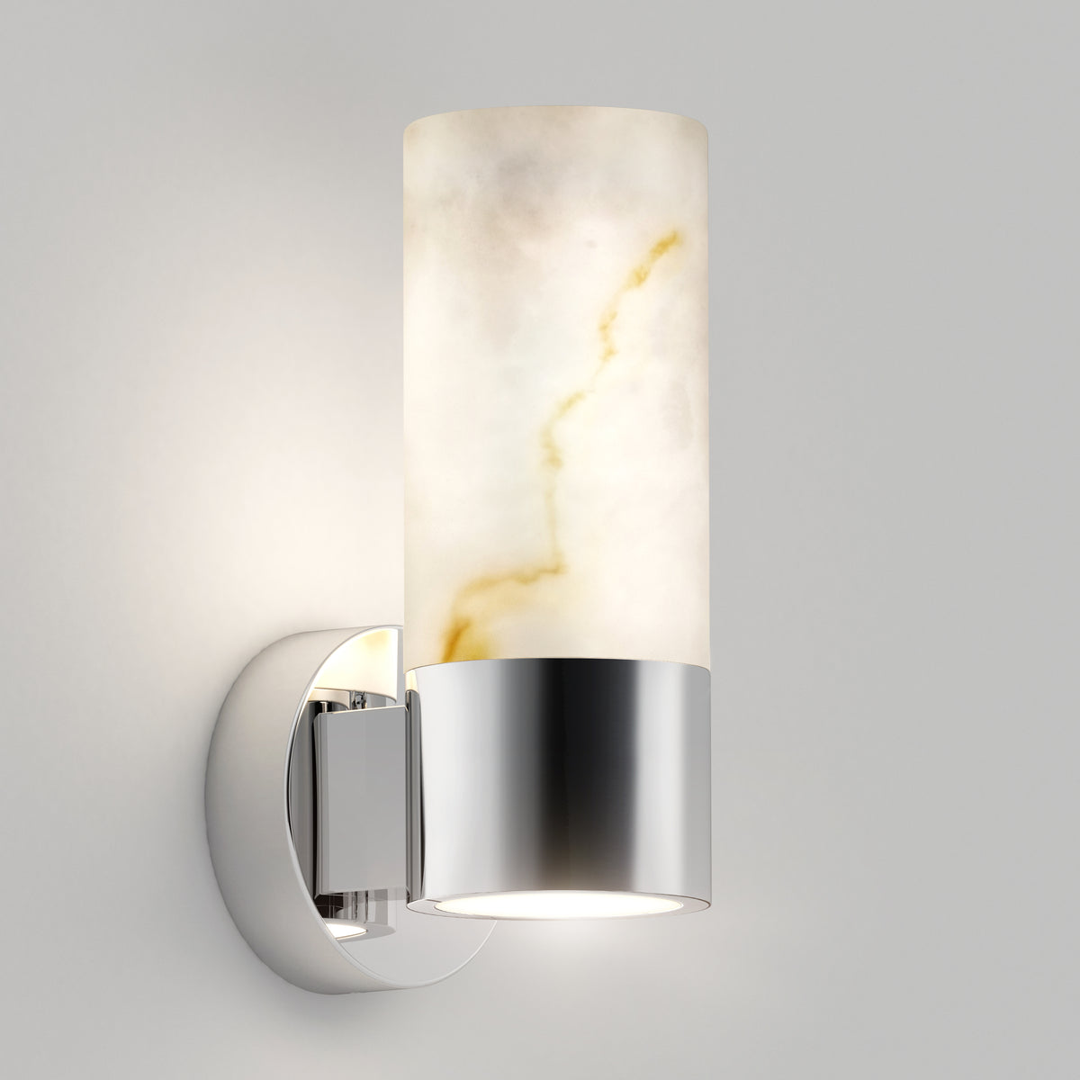 A modern wall sconce with a cylindrical frosted glass shade and a polished chrome base, mounted on a white wall. The light glows softly through the glass, showcasing subtle marbled patterns.