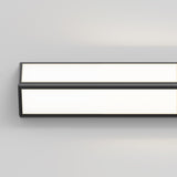 A modern, rectangular wall-mounted LED light fixture with a black frame, glowing with a bright white light against a plain light gray wall.