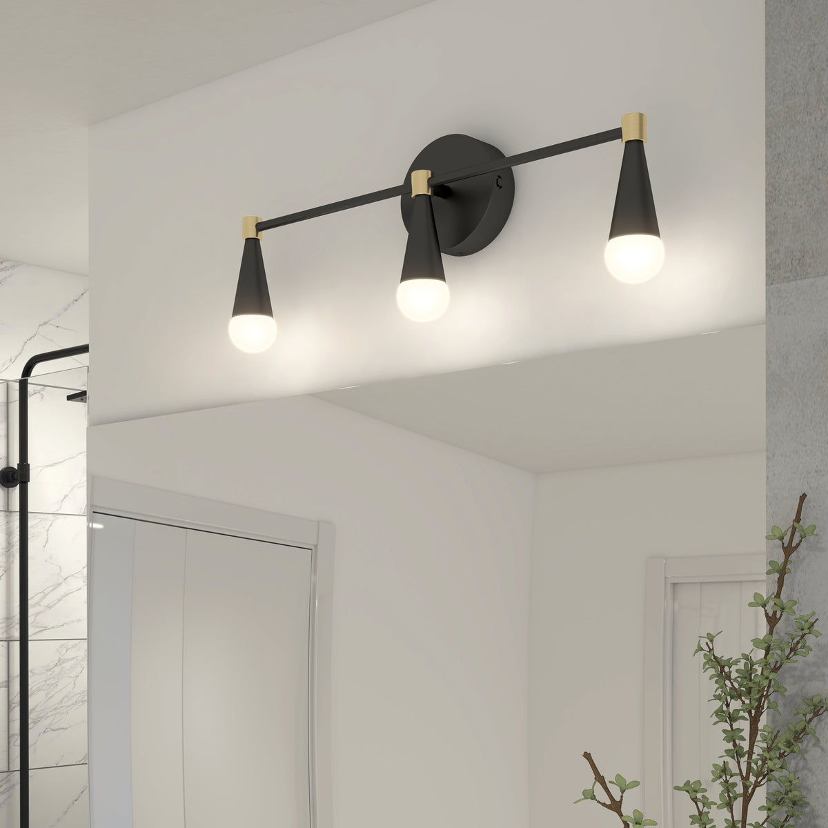 A modern bathroom light fixture with three black and gold cone-shaped lights is mounted above a mirror, illuminating the white wall and reflecting in the glass. A shower and a plant are partially visible nearby.