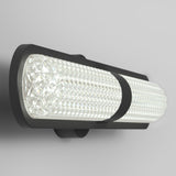 A modern wall-mounted light fixture with a cylindrical, textured glass shade and a sleek black metal frame, emitting bright, even illumination against a light gray wall.
