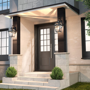 Modern front entrance with a black door featuring glass panes, two Artika Colonial outdoor lantern wall lights on either side, stone steps, and small green shrubs flanking the entryway.