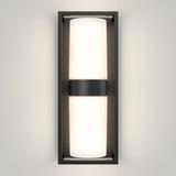 A modern wall sconce with a rectangular black frame and a central horizontal black band, featuring a frosted white cylindrical light emitting a soft glow against a light-colored wall.