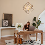 A woman sits at a wooden desk in a stylish, modern home office, writing in a notebook beside a laptop, a vase of white flowers, and décor, with plants and artwork in the background.