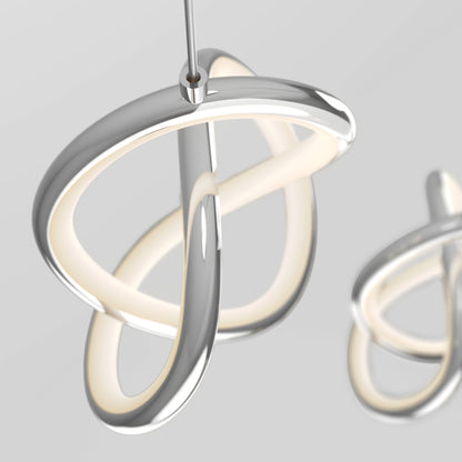 A modern, sculptural LED pendant light with smooth, intertwined silver loops and a soft inner glow, hanging from a thin cable against a neutral gray background. Another similar light is blurred in the background.