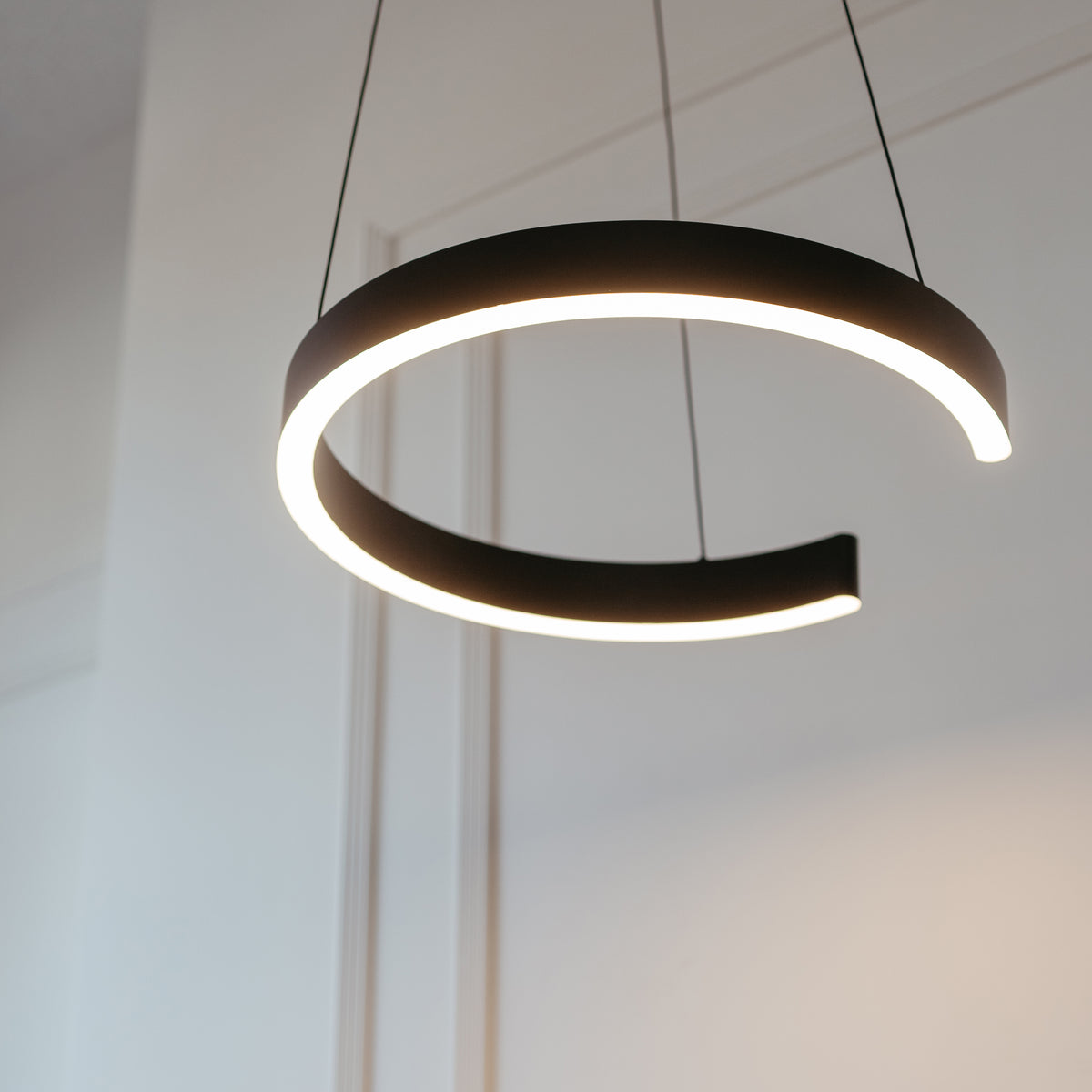 A modern, circular pendant light fixture with a gap, emitting a soft white glow, hangs from the ceiling by two thin wires in a minimalistic room with white walls.