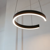 A modern, circular pendant light fixture with a gap, emitting a soft white glow, hangs from the ceiling by two thin wires in a minimalistic room with white walls.