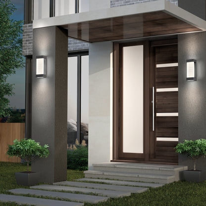 Modern house entrance with a wooden front door featuring horizontal glass panels, Percy LED indoor-outdoor wall lights by Artika on each side, potted plants flanking the entry, and concrete stepping stones leading to the doorway.