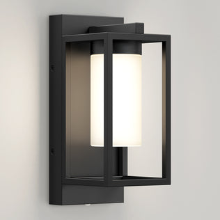 The Artika Aurel Outdoor LED Light, 3CCT features a modern black rectangular frame with a cylindrical white frosted glass shade, mounted on a light wall and emitting a soft, warm glow.