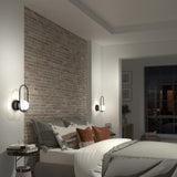 A modern bedroom with a brick accent wall, neatly made bed with layered pillows, round bedside tables, wall-mounted globe sconces, and a window showing a nighttime city view.
