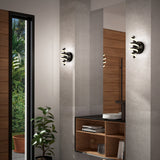 A modern interior with gray walls, two sculptural black and gold wall sconces, a wooden shelving unit with books and decor, a round white sink, and a potted plant near a tall window with a view of greenery.