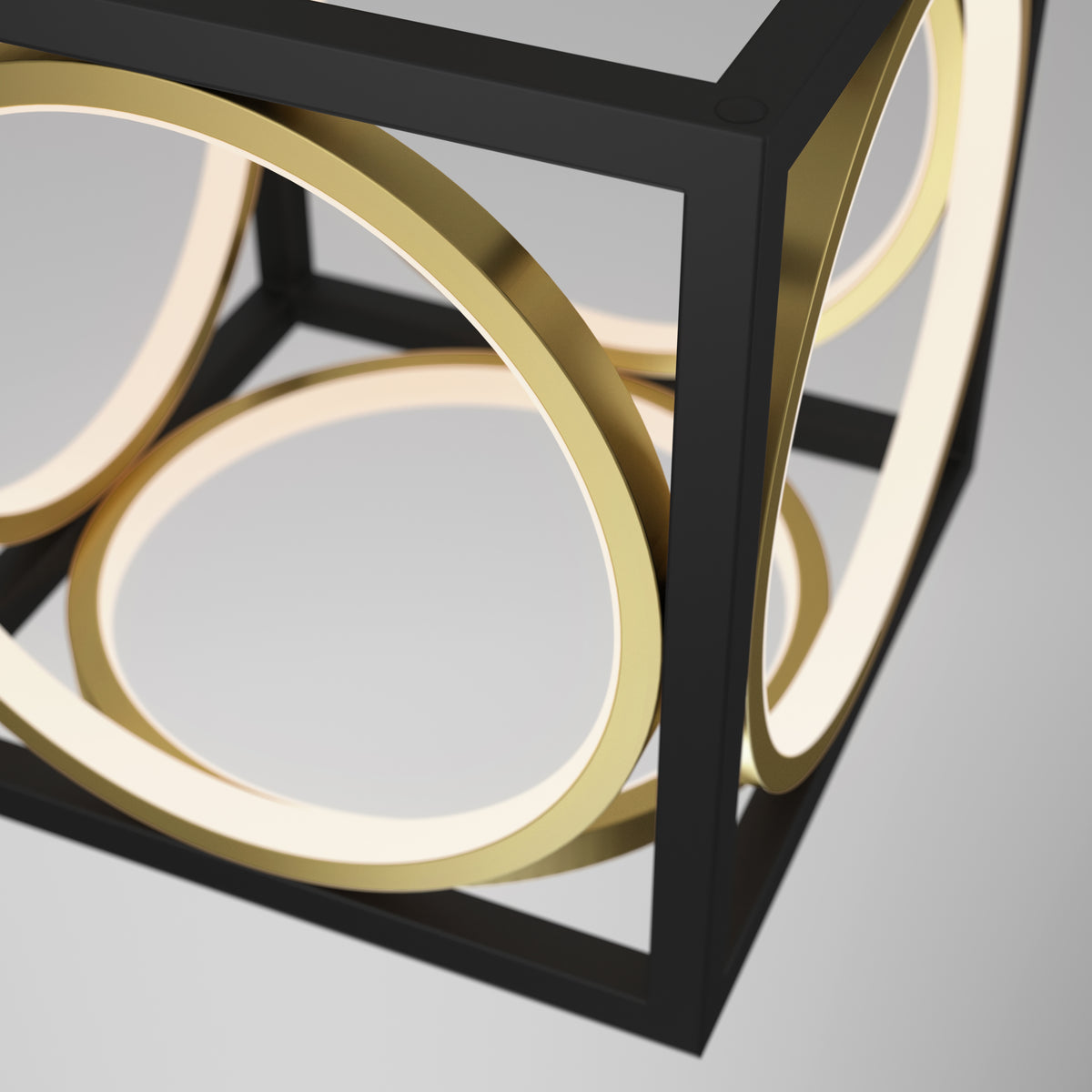A modern, geometric light fixture featuring a black open cube frame with intersecting gold rings that emit a soft white light, suspended against a neutral gray background.
