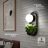 A modern wall sconce with a matte black finish features a glowing glass diffuser and a built-in planter with green foliage, mounted on a white brick wall beside framed abstract art. Text highlights LED, glass diffuser, and 3-year warranty.