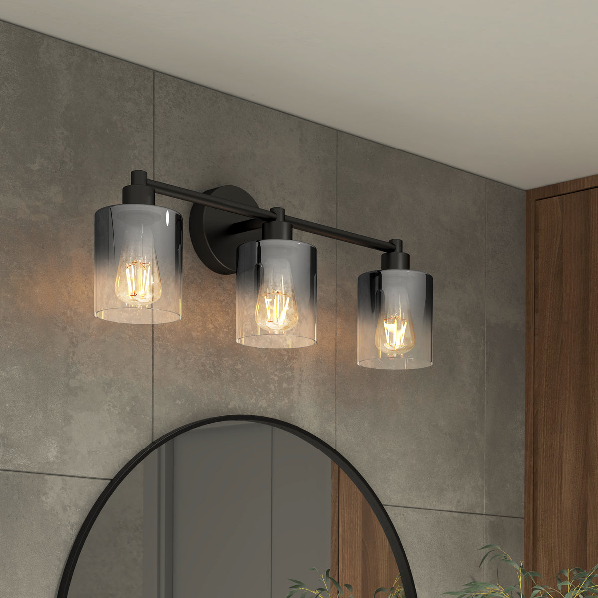 Three-bulb wall sconce with cylindrical glass shades and exposed filament bulbs mounted above a round mirror on a gray tiled wall, with a wooden accent panel and green plant visible in the scene.