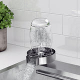 A glass is placed upside down on a modern glass rinser installed in a kitchen sink, with water spraying up inside the glass. There is a potted plant nearby and a white tiled backsplash in the background.