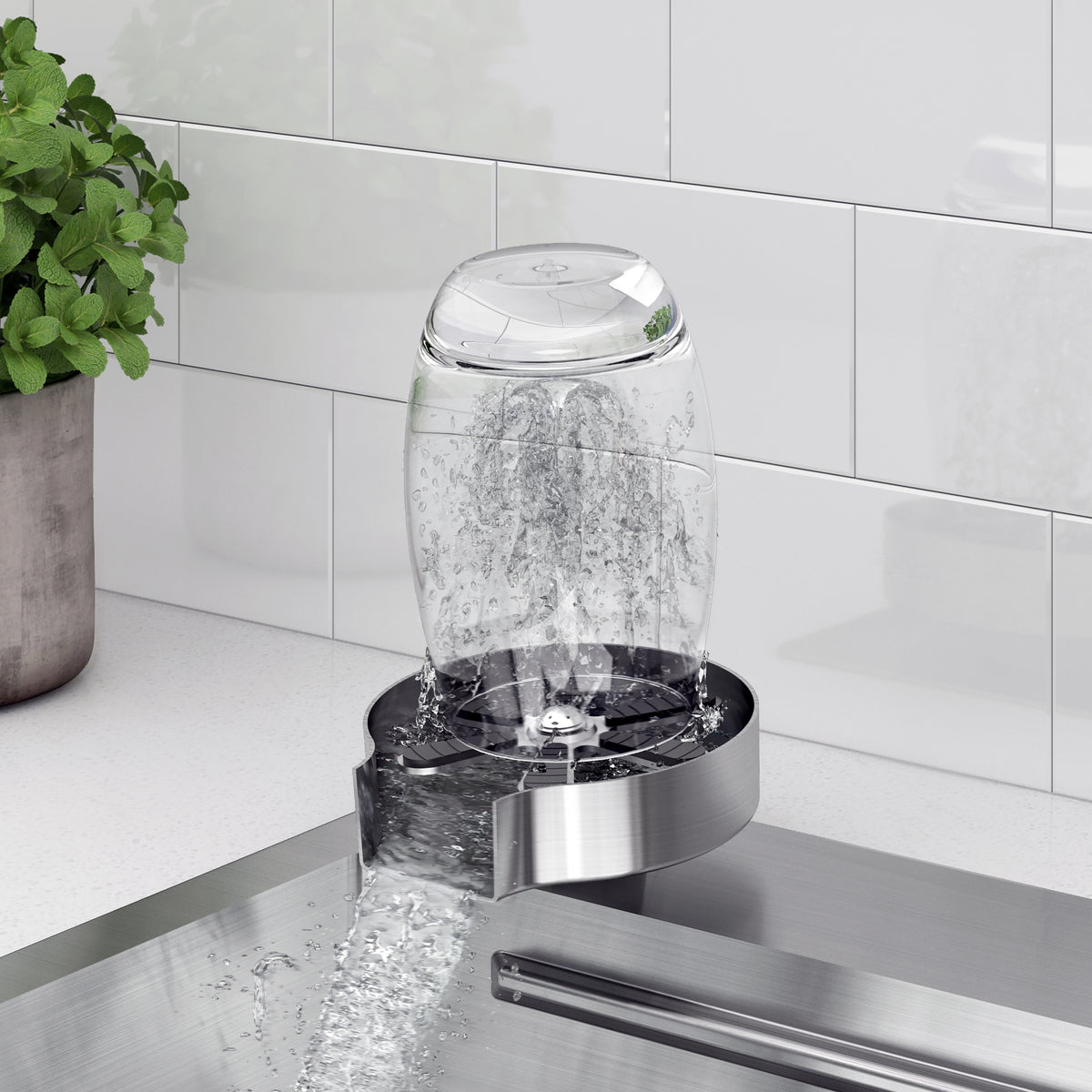 A glass cup is placed upside down on a glass rinser beside the Artika Neoflow 31 in. stainless steel workstation sink, with water spraying upwards to clean the inside, next to a potted plant and white tiled backsplash.