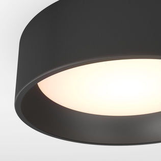 A close-up of the Artika Alton LED flush mount features a sleek black circular shade and glowing white light, set against a plain white ceiling.
