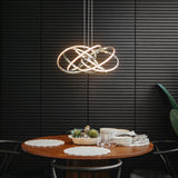 A modern dining area with a round wooden table set for four, featuring placemats, plates, and glasses. Above the table hangs a contemporary LED pendant light with interlocking rings, against a dark, textured wall.