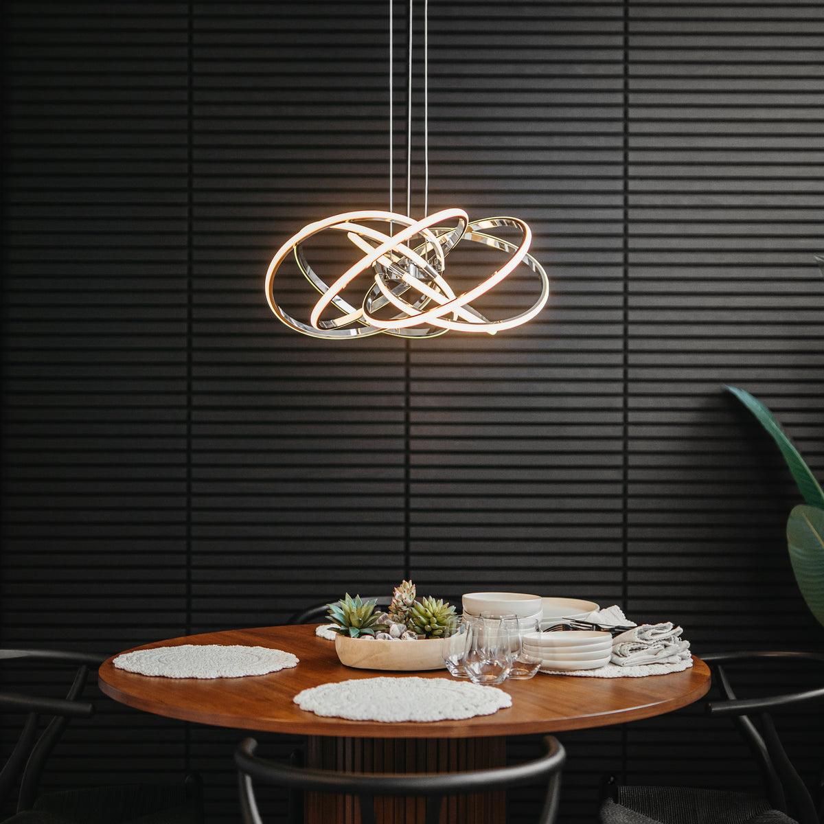 A modern dining area with a round wooden table set for two is illuminated by the Artika Maelstrom modern LED pendant light 5 CCT, hanging above. The scene features white placemats, dishes, glassware, and a black textured wall.