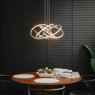 A modern dining area with a round wooden table set for two is illuminated by the Artika Maelstrom modern LED pendant light 5 CCT, hanging above. The scene features white placemats, dishes, glassware, and a black textured wall.