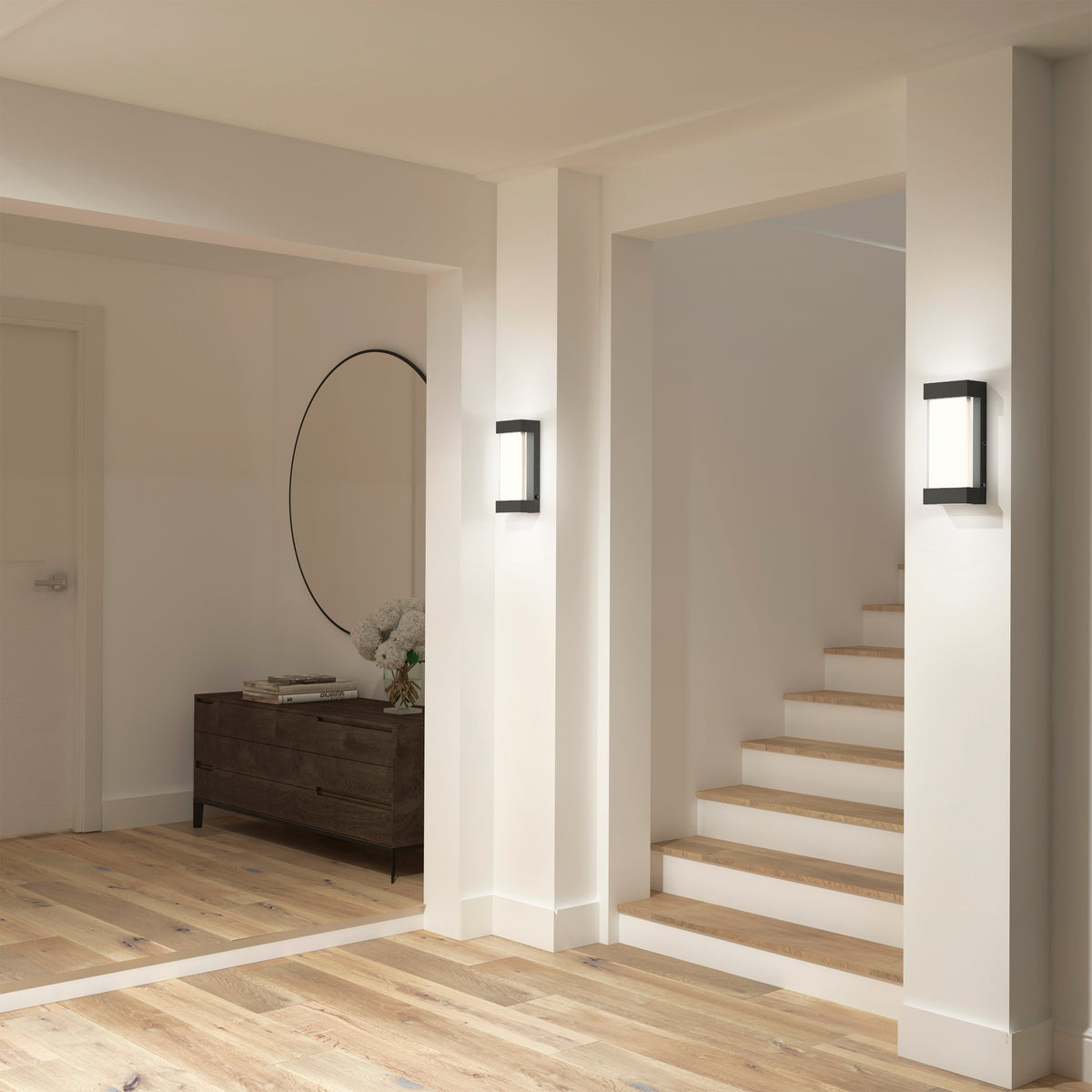 A modern hallway with light wood floors, white walls, a staircase, a round mirror above a dark wood console, white flowers, and two Artika Glacier indoor-outdoor LED wall lights.