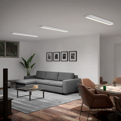 A modern living room features a gray sofa, glass coffee table, large plant, framed wall photos, and a dining area with brown leather chairs and a round table—all illuminated by the Artika Aiden 48 in. LED modern flush mount light.