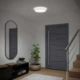 A modern entryway with a gray door, a round ceiling light, a potted plant, a black bench with brown cushions, an oval wall mirror, and wooden stairs with a metal railing.