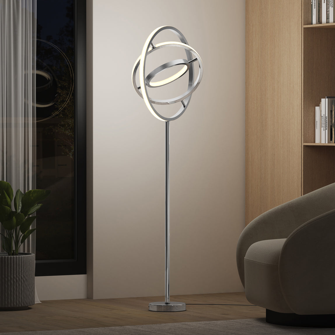 A modern floor lamp with circular, orbit-like LED rings stands on a metal base beside a beige armchair, wooden bookshelf, and potted plant in a contemporary living room.