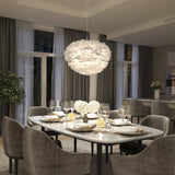 A modern dining room with a marble table set for six, featuring elegant dishware and white floral centerpieces. Plush gray chairs surround the table, and a large, feathery pendant light hangs above. Floor-to-ceiling curtains line the windows.