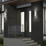 Modern front entrance with dark brick pillars, white frosted glass double doors, metal railing, and wall-mounted cylindrical lights on each side of the door, illuminated at twilight. Concrete steps lead to the entryway.