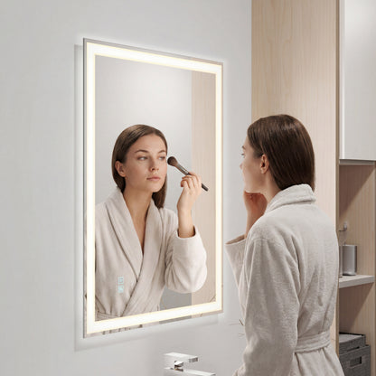 A woman in a white bathrobe applies makeup with a brush, looking into the Artika Sofia LED mirror 3 CCT with defogger. The bathroom showcases light wood cabinets and minimalistic decor.