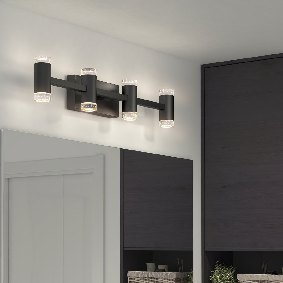 A modern black vanity light fixture with four cylindrical LED lights mounted above a bathroom mirror, illuminating the space. Dark cabinets and part of a white door are visible in the reflection.