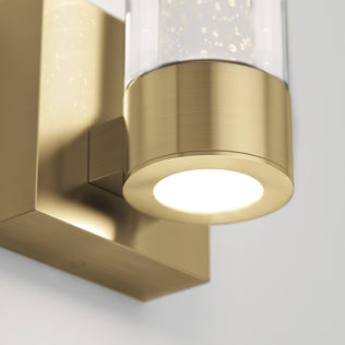 Close-up of Artika's Essence 1-light LED bathroom vanity light with a gold finish and a cylindrical glass shade on a white wall.