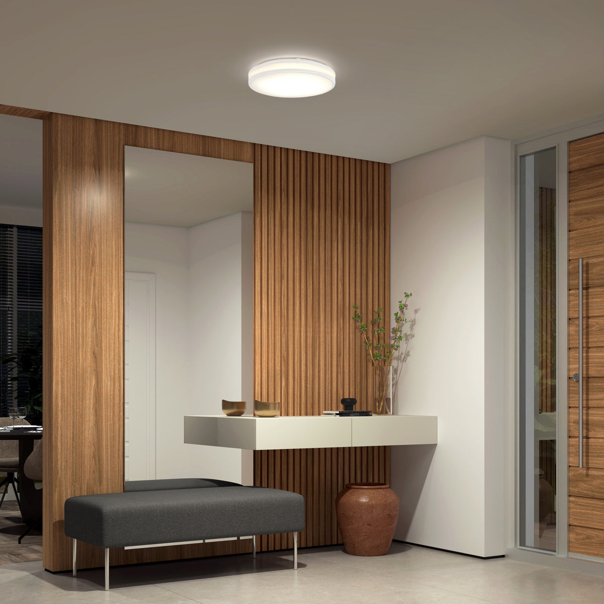 A modern entryway features wood panel walls, a large mirror, a floating white shelf with decor, a dark cushioned bench, and a ceramic vase. The Artika Hemisphere 15 in. LED flush mount light offers adjustable brightness for a stylish touch.