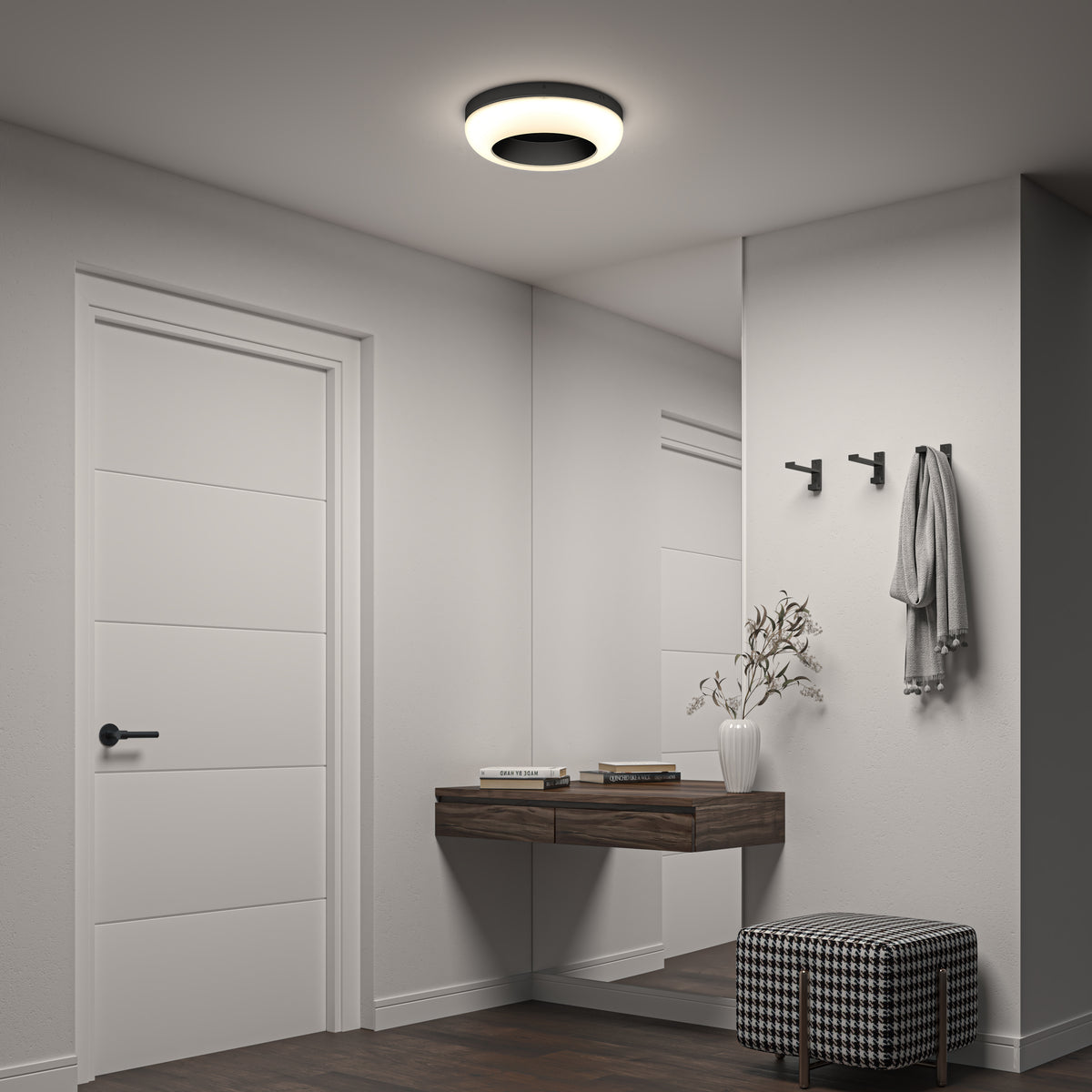 A modern, minimalist hallway features a white door, a wall mirror, a floating wooden shelf with books and a white vase, two black wall hooks, and a checkered cushioned stool under a circular ceiling light.