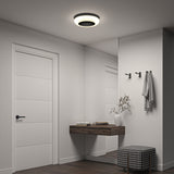 A modern, minimalist hallway features a white door, a wall mirror, a floating wooden shelf with books and a white vase, two black wall hooks, and a checkered cushioned stool under a circular ceiling light.