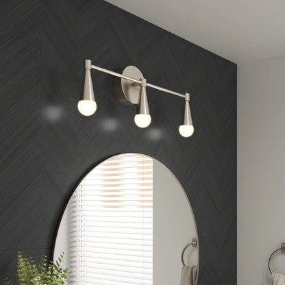 The Artika Wayden LED mid-century bathroom vanity light 3 CCT with three glowing bulbs is mounted above a round mirror on a dark chevron-patterned wall, alongside a small plant and towel ring.