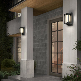 A modern home entrance features gray stone walls, a glass-paneled door, concrete pillars under a wooden ceiling, and two Artika Aurel Outdoor LED Light 3CCT wall fixtures. Steps can be seen inside through the door.