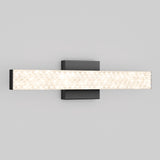 A modern wall-mounted light fixture with a rectangular, textured illuminated bar extending horizontally from a square, matte black base against a plain light gray background.