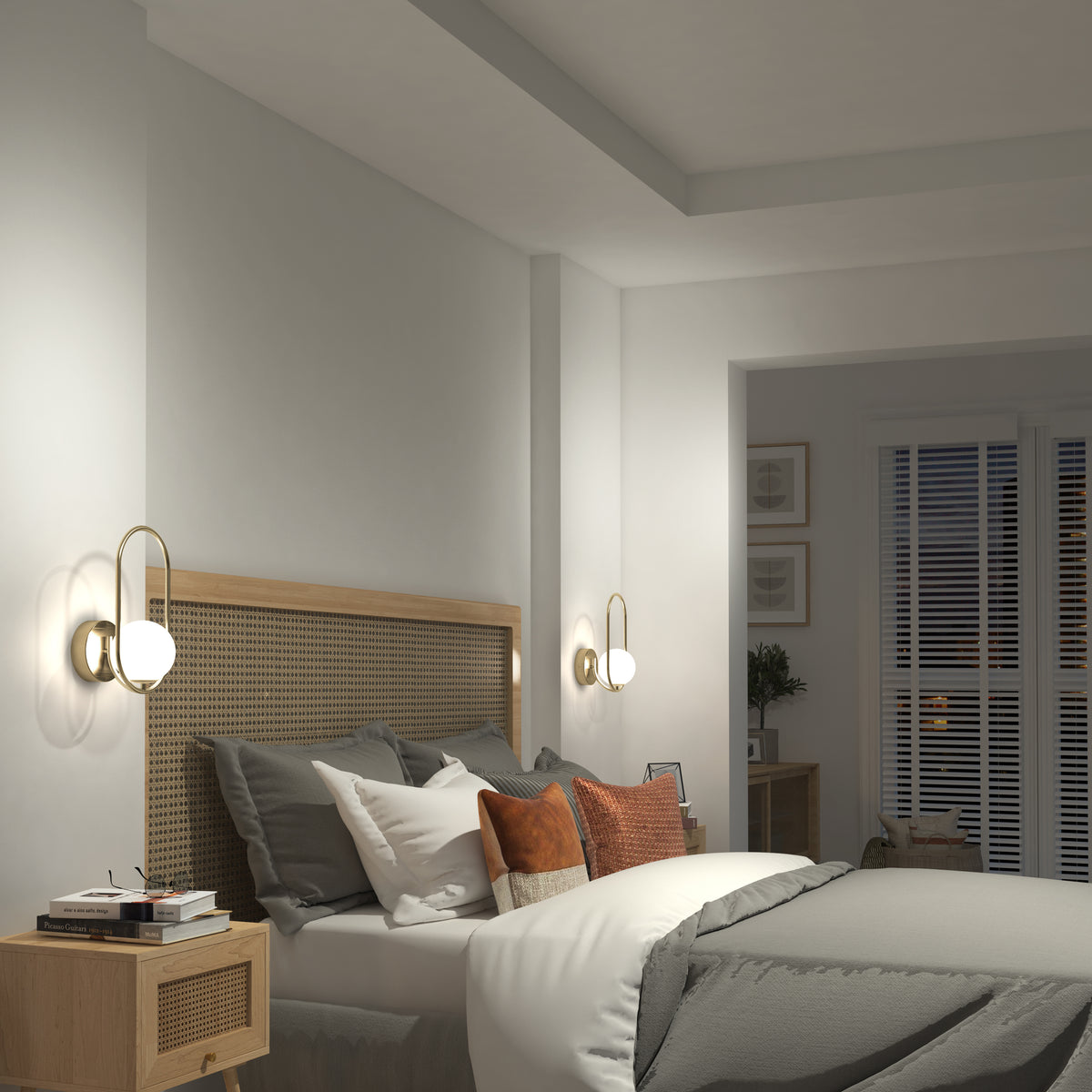 A cozy bedroom features a wicker headboard, gray bedding, and accent pillows. Two Artika Bloom LED Sconce 3 CCT lights offer soft illumination. A nightstand holds a book, and window blinds let in natural light.