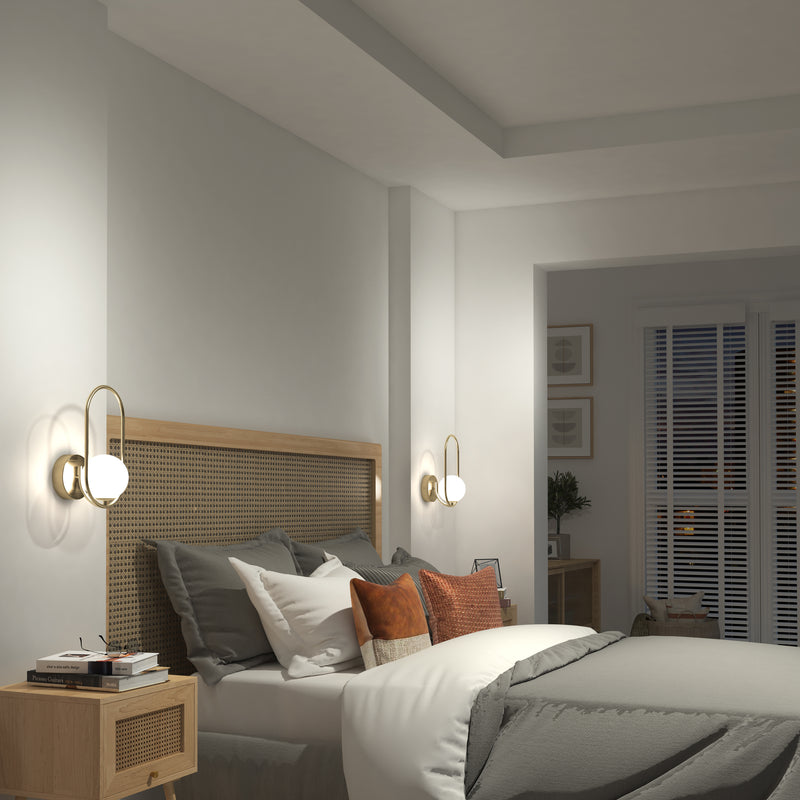 A cozy bedroom features a wicker headboard, gray bedding, and accent pillows. Two Artika Bloom LED Sconce 3 CCT lights offer soft illumination. A nightstand holds a book, and window blinds let in natural light.