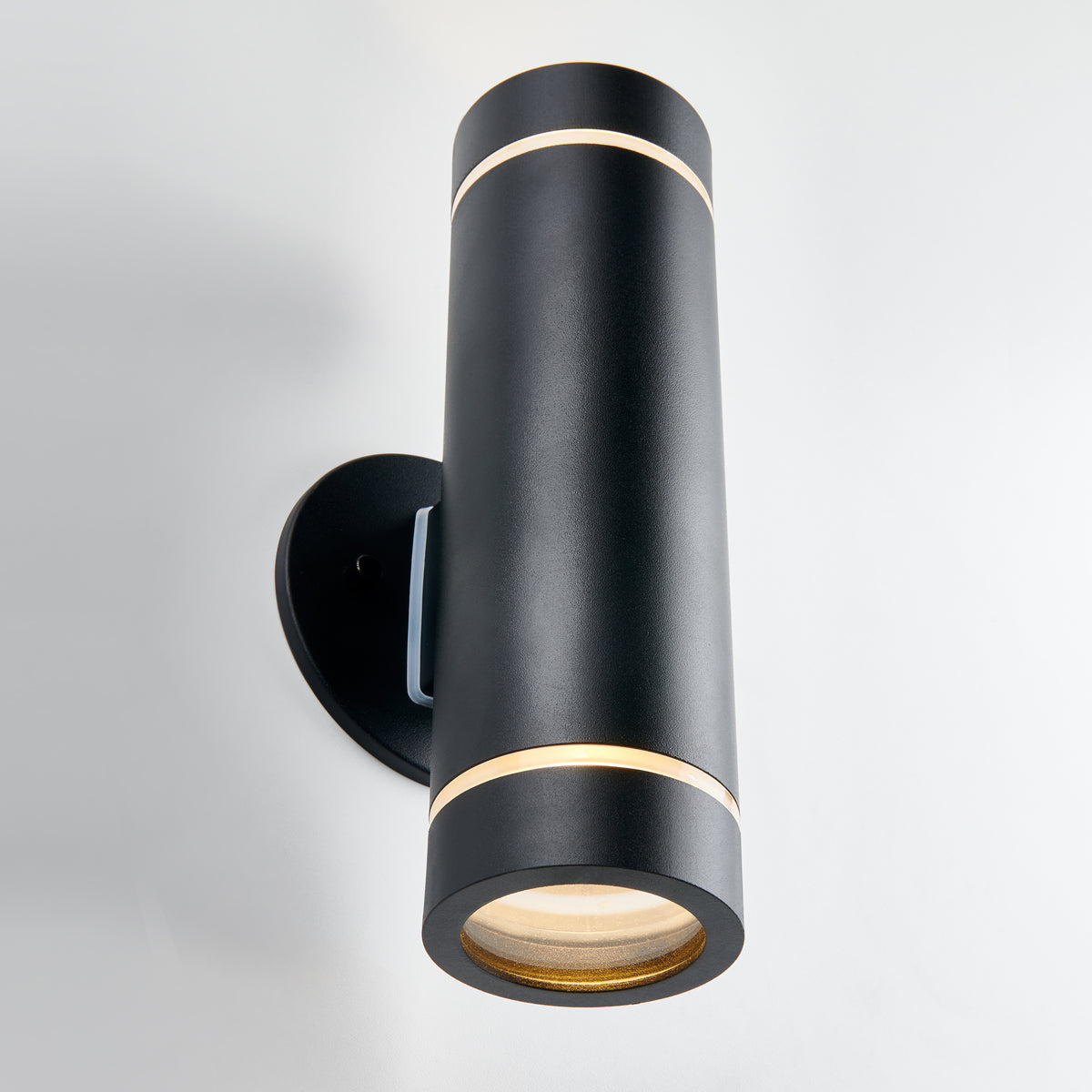 A modern black cylindrical wall sconce with thin gold accents, mounted vertically on a white wall, emitting light from both the top and bottom ends.