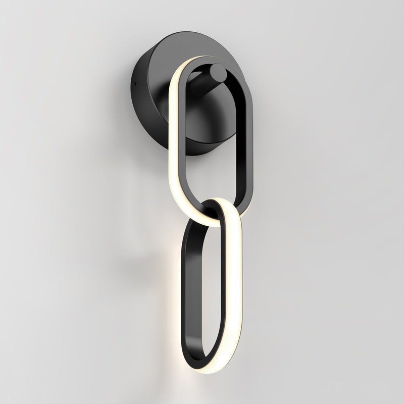 The Artika Catena modern LED wall sconce 5 CCT features two interlocking oval loops—one illuminated, one solid black—mounted on a round black base against a white wall.